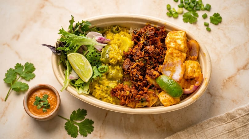 A colorful Indian food bowl with dal, paneer, and a side of sauce on a marble surface.