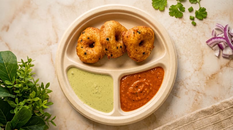 Three golden-brown medu vada on a segmented plate with green and red chutneys, garnished with herbs and onions.